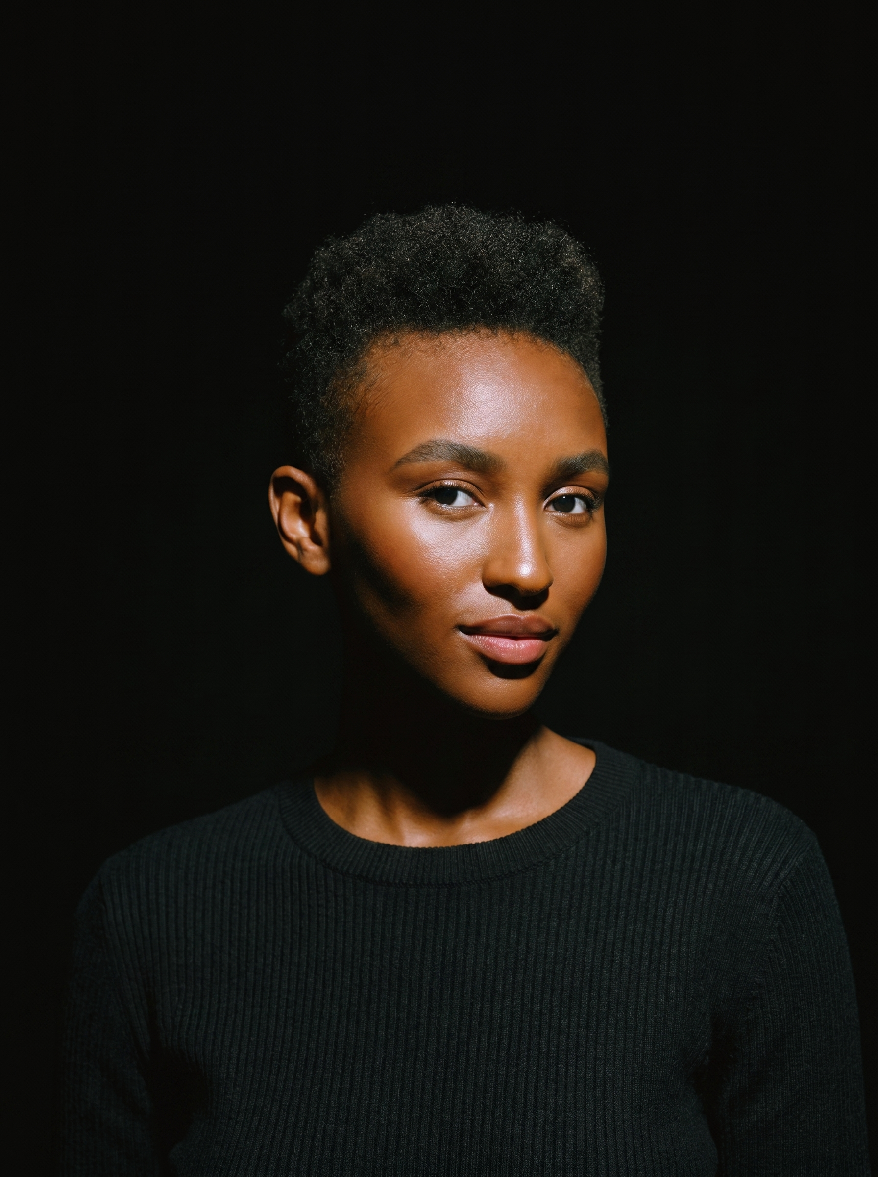 Tight close portrait, black ribbed sweater, single hard key light, deep black void.